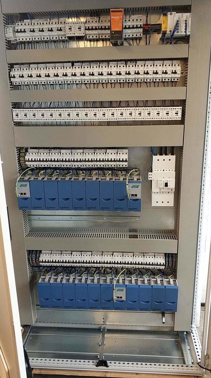 Otwarta szafa sterownicza z licznymi elementami elektrycznymi, w tym zabezpieczeniami, modułami sterującymi i okablowaniem, ułożonymi w regularny sposób.