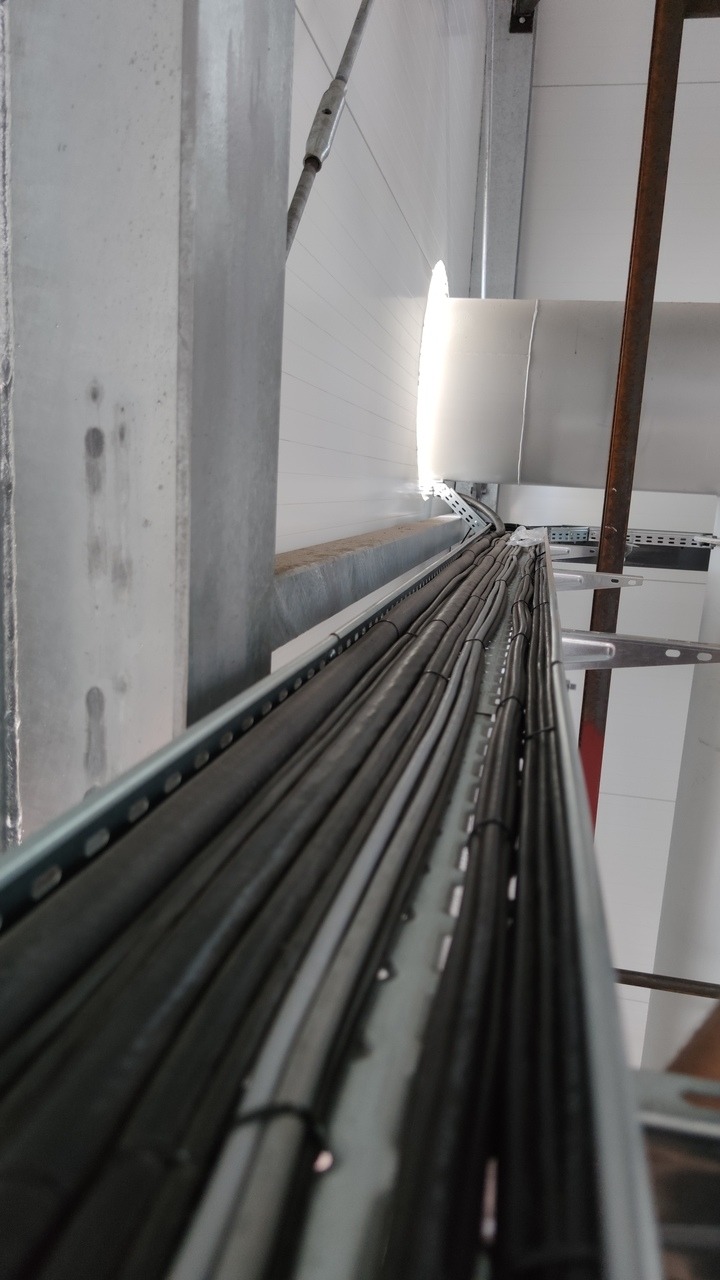 Metalowa korytka kablowe wypełnione przewodami elektrycznymi, zamocowane na konstrukcji wsporczej wewnątrz budynku przemysłowego, widok z dołu.