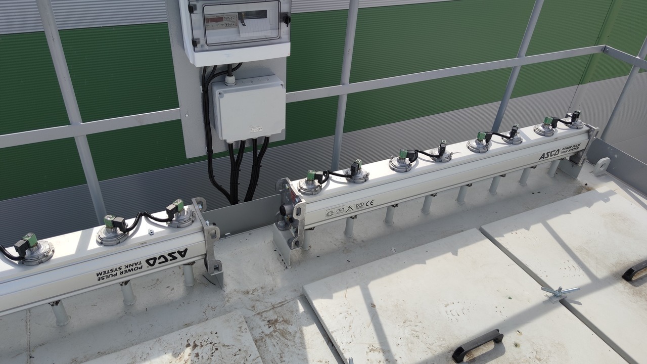 Widok z góry na zainstalowany system ASCO Power Pulse Tank System z czujnikami, zamontowany na białej powierzchni, z szarą metalową konstrukcją wsporczą i zielono-szarą elewacją budynku w tle.