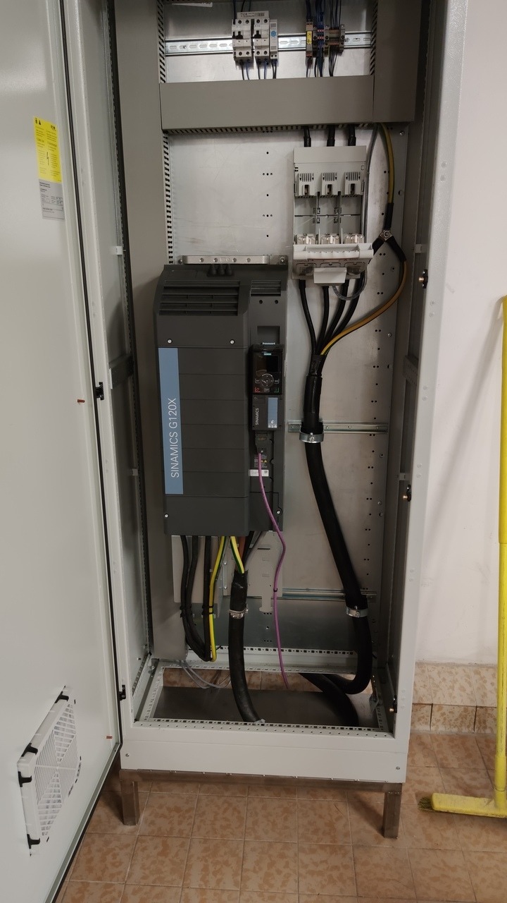 Otwarta szafa sterownicza z zamontowanym falownikiem Siemens Sinamics G120X i okablowaniem, widoczne zabezpieczenia nad falownikiem i wentylator w drzwiach.