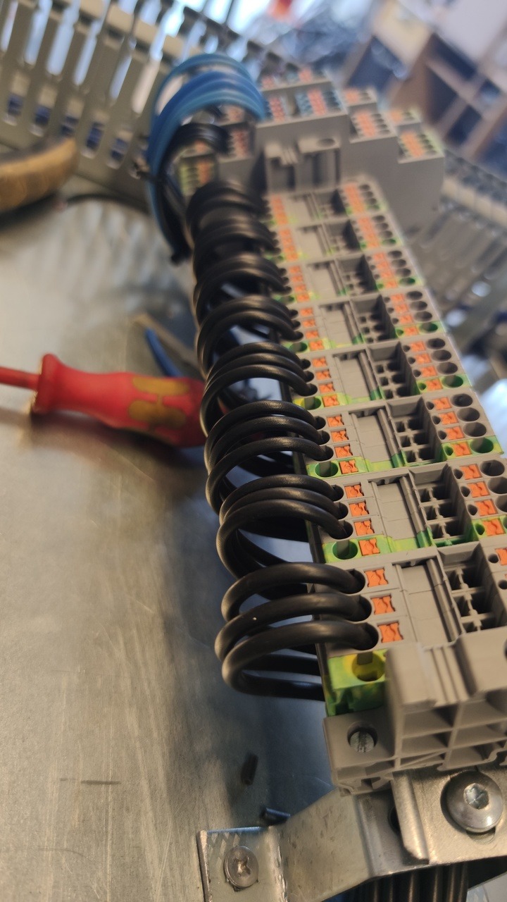 Szyna DIN z podłączonymi przewodami w izolacji spiralnej, widoczne złącza elektryczne i częściowo czerwony śrubokręt na metalowym blacie w tle.