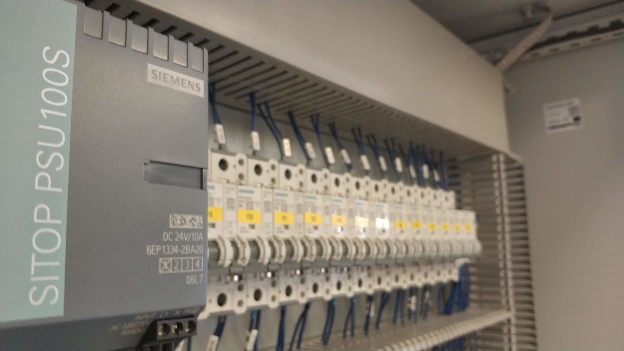 Szary zasilacz Siemens SITOP PSU100S w szafie sterowniczej z rzędami zabezpieczeń nadprądowych, podłączonych niebieskimi przewodami.