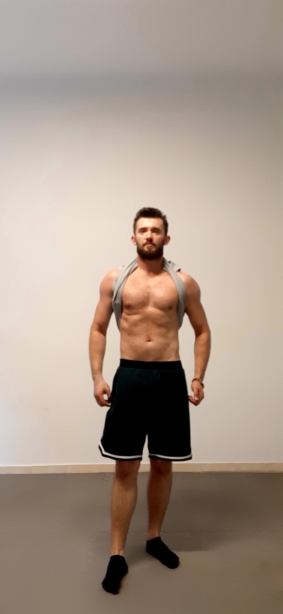 Mężczyzna z brodą, w sportowych szortach i skarpetkach, z koszulką przerzuconą przez ramiona, prezentuje wyrzeźbioną sylwetkę na tle jasnej ściany.