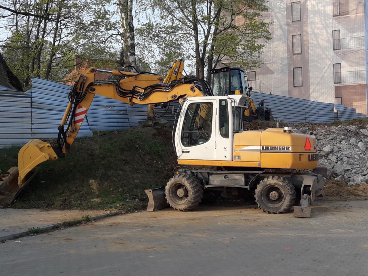 Żółto-biała koparka kołowa Liebherr na placu budowy, widoczna z boku, z fragmentem wykopu i ogrodzeniem w tle.