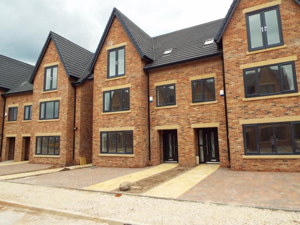 Nowe szeregowe domy z cegły z ciemną stolarką okienną i brukowanym podjazdem z jasnymi pasami, widok z poziomu gruntu.