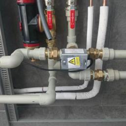 LepszePowietrze.com - Węzeł instalacyjny z zaworami, manometrem, filtrem i przewodami z tworzywa sztucznego oraz przewodami miedzianymi w izolacji, zamontowany na szarej ścianie z drzwiczkami rewizyjnymi z kluczem.