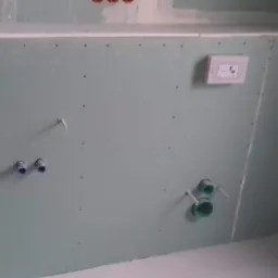 Ściana z płyty gipsowo-kartonowej w trakcie montażu, widoczne przyłącza wodne i elektryczne, oraz otwór rewizyjny