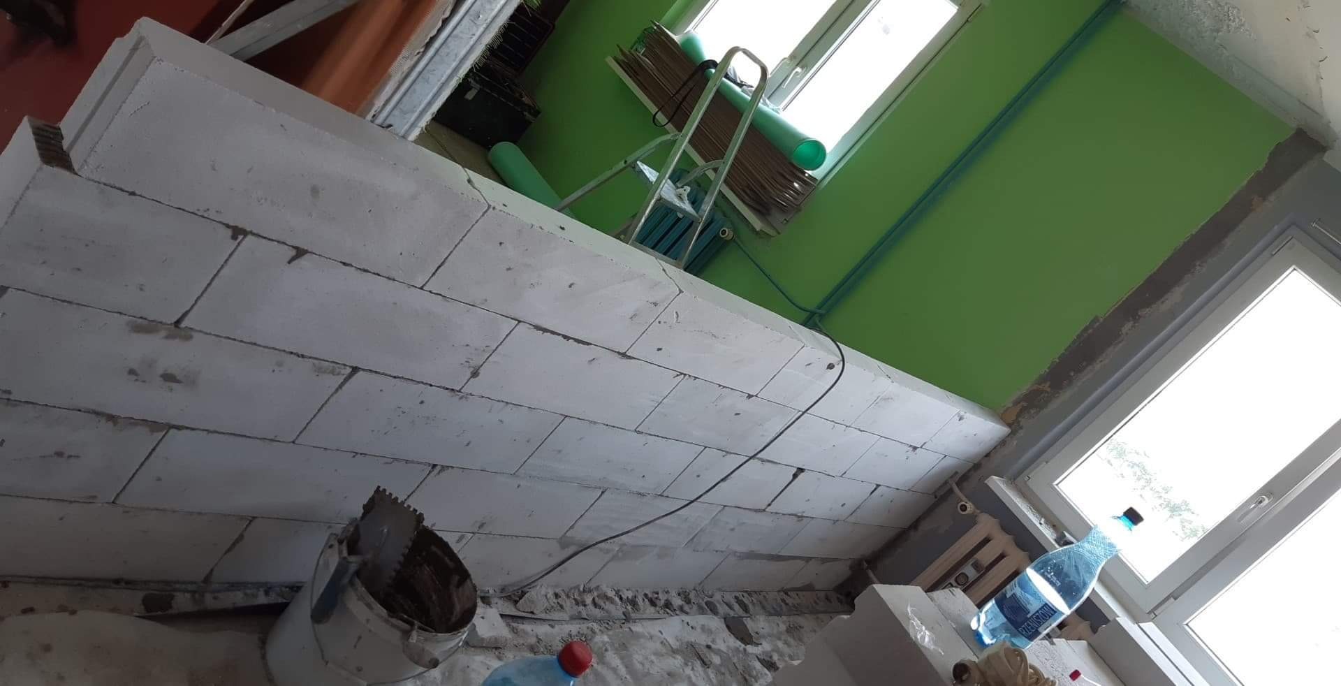 Wznoszenie ściany z bloczków betonowych, widoczny fragment ściany, drabina oparta o ścianę z zieloną rurą, wiadro z narzędziami, butelka wody.