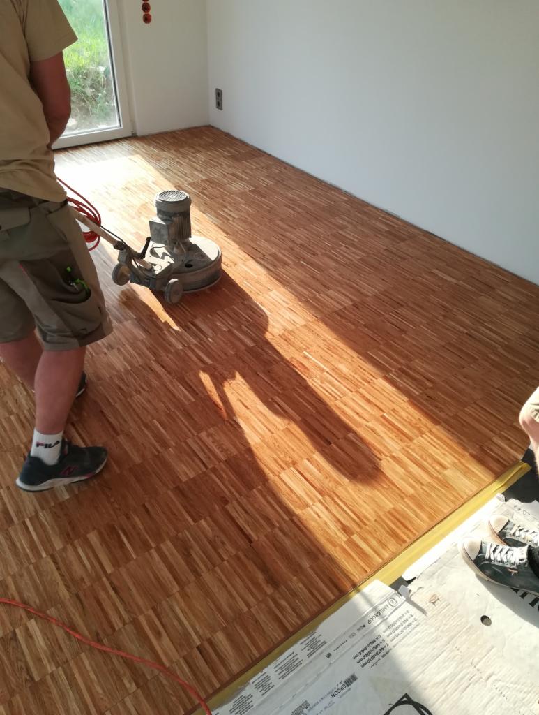 Maszyna szlifująca drewnianą podłogę parkietową w jasnym pomieszczeniu, widoczne częściowo dwie osoby i opakowania ochronne na podłodze.