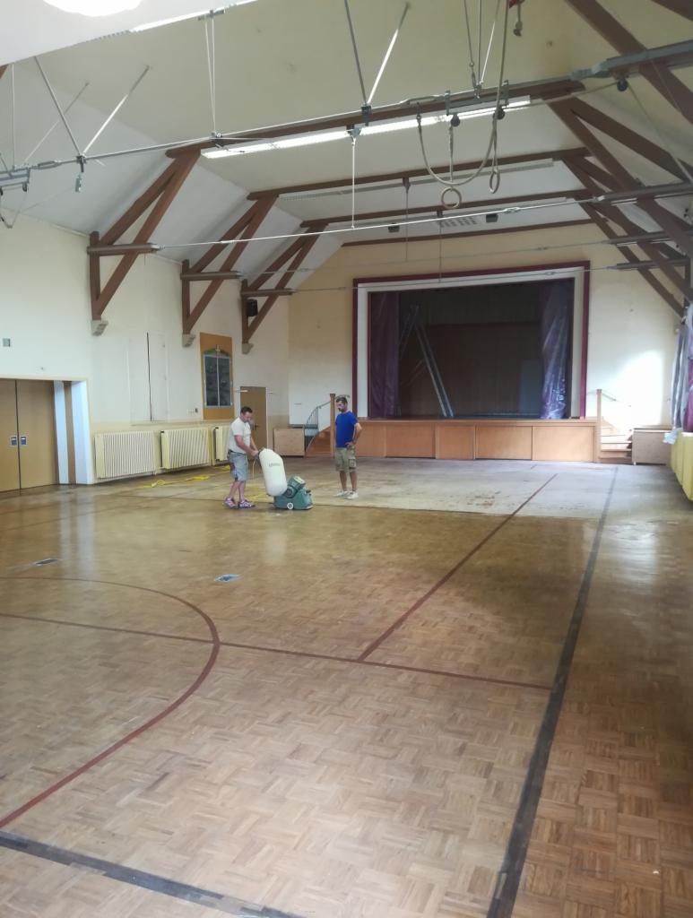 Sala gimnastyczna z drewnianą podłogą, na której dwóch mężczyzn używa maszyny do polerowania parkietu, widoczna scena w tle.