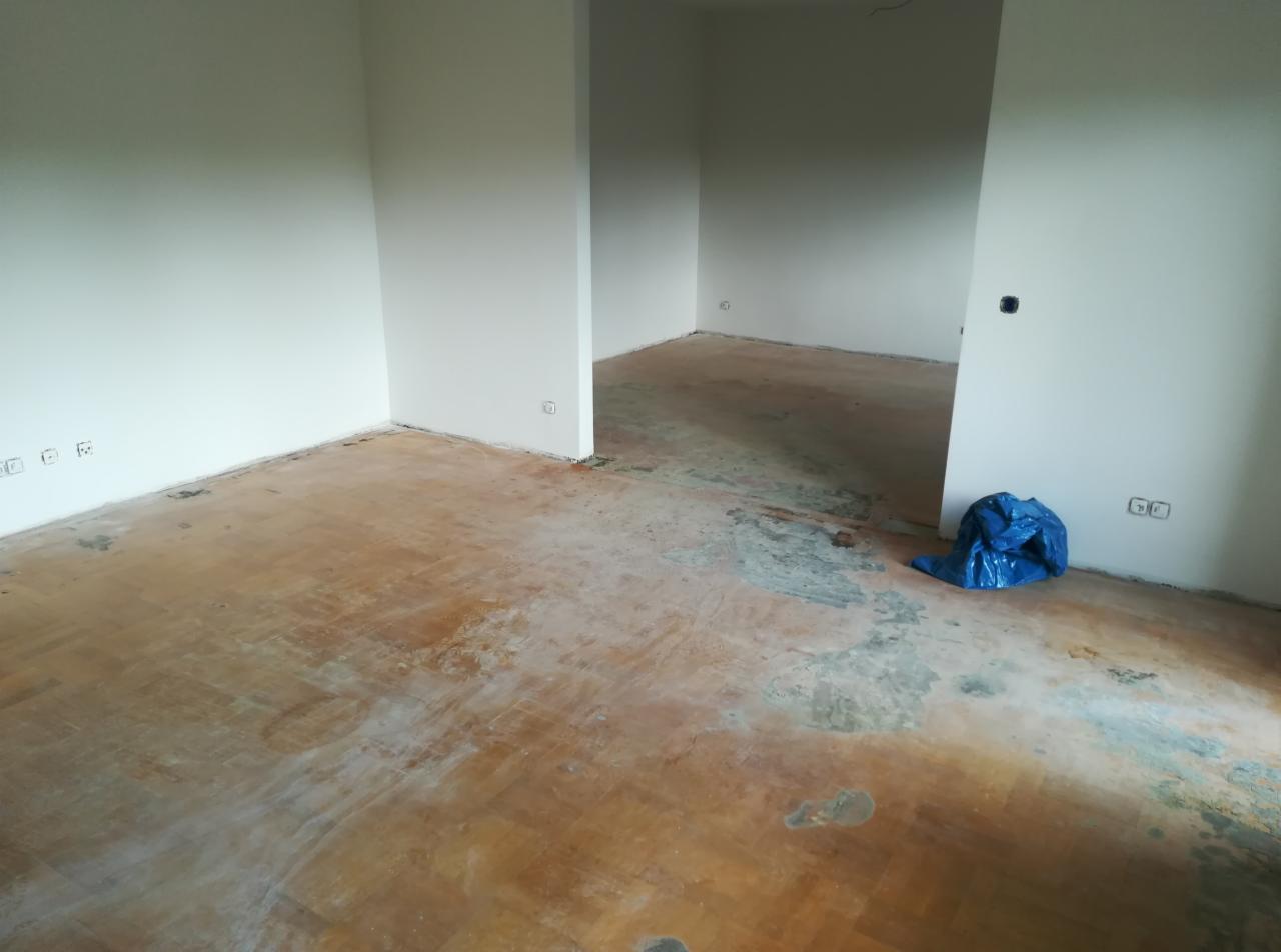 Pokój w trakcie remontu: stara, zniszczona drewniana podłoga z pozostałościami po kleju, białe ściany, niebieski worek na podłodze, widoczne gniazdka elektryczne.