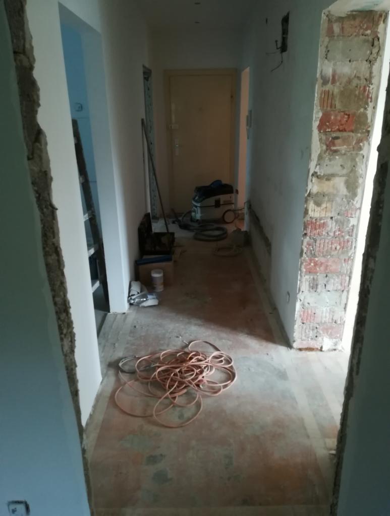 Korytarz w trakcie remontu: widoczne odsłonięte cegły, przewody na podłodze, drabina w tle i odkurzacz przemysłowy, sugerujące prace wykończeniowe.