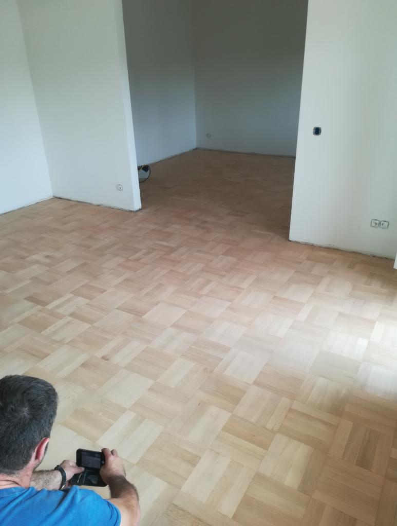 Jasna, nowo ułożona podłoga z parkietu w jodełkę w pustym, odświeżonym pokoju. Mężczyzna fotografuje podłogę telefonem, widoczny z tyłu, ubrany w niebieską koszulkę.