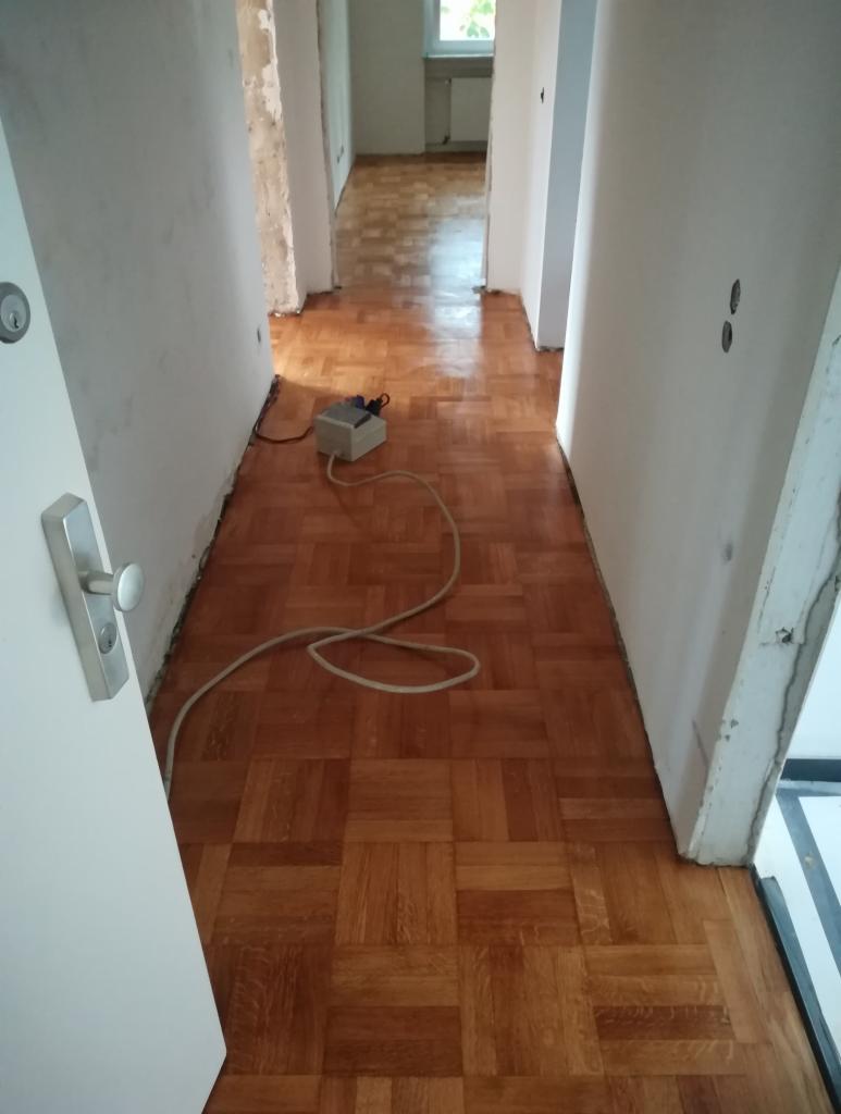 Korytarz z ułożonym parkietem w kwadraty, widoczny kabel i gniazdko elektryczne na podłodze, ściany w trakcie remontu.