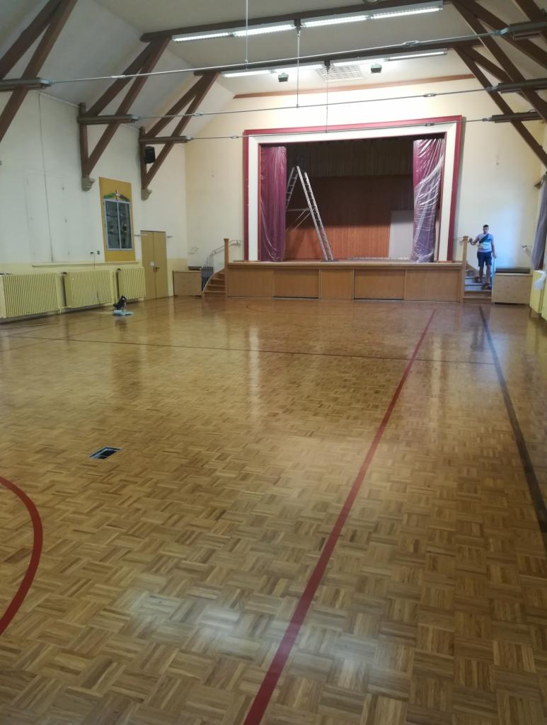 Sala gimnastyczna z parkietem w jodełkę po renowacji, widoczna scena z drabiną i pracownik stojący na schodach, parkiet świeżo polakierowany z liniami boiska.