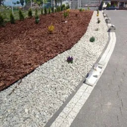 Aranżacja ogrodu z korą, kamieniem ozdobnym i niskimi roślinami, oddzielona obrzeżem od kostki brukowej.