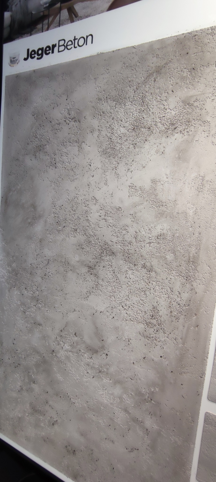 Szara, teksturowana ściana z efektem betonu, oświetlona z góry. Widoczny fragment logo 'Jeger Beton' w lewym górnym rogu. Delikatne cieniowanie podkreśla nierówności powierzchni.