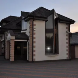 Dwupiętrowy dom z ciemnobrązowym dachem, jasną elewacją, ceglanymi narożnikami i dużym oknem, widok z zewnątrz na podjeździe z kostki brukowej.