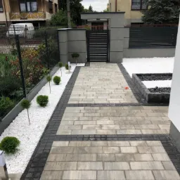 Ułożona kostka brukowa w odcieniach szarości prowadząca do furtki z metalowymi sztachetami, obok rabata z białym kamieniem i małymi kulistymi krzewami.