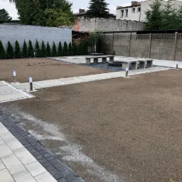 Przygotowany teren pod trawnik z wytyczonymi ścieżkami z płyt betonowych i białego grysu, betonowymi ławkami i oświetleniem ogrodowym, w tle wysokie tuje i betonowy płot.