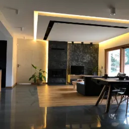 Nowoczesny salon z podświetlanym sufitem, ścianą z ciemnego kamienia za kominkiem, betonową posadzką przechodzącą w drewnianą, oraz czarnym stołem z krzesłami w tle okna.