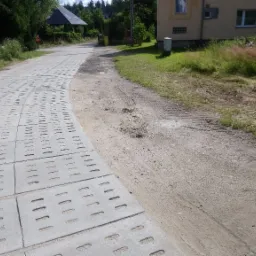 Ułożona droga z betonowych płyt ażurowych, widoczne ślady opon na poboczu, w tle budynki mieszkalne i zieleń.