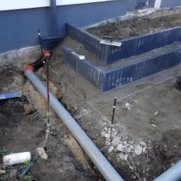 Szara rura kanalizacyjna z pomarańczowymi złączkami w wykopie obok fundamentów schodów z kostki, widoczne sznurki geodezyjne i narzędzia murarskie.