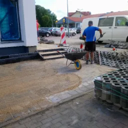 Układanie kostki ażurowej na posesji: robotnik z łopatą, taczka z kostką, przygotowane podłoże z piasku, kostka ażurowa w paletach, budynek z oknami zabezpieczonymi folią w tle.