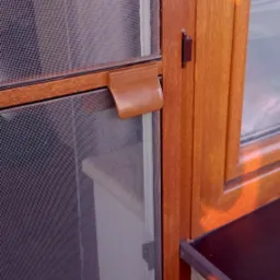 Zamknięta moskitiera ramkowa w oknie z brązowego PCV, widoczny uchwyt do przesuwania siatki i fragment muru z parapetem.