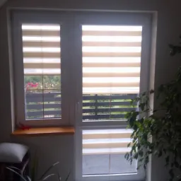 Dwuokienne okno na poddaszu z zamontowanymi roletami w odcieniach beżu z poziomymi pasami, widok na balkon i zieleń, po prawej stronie duża roślina doniczkowa.