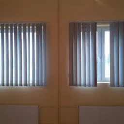 Dwa okna z szarymi żaluzjami pionowymi na żółtej ścianie, jedno okno z żaluzjami częściowo otwartymi, widoczne grzejniki pod oknami.