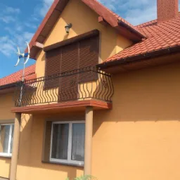 Brązowe rolety zewnętrzne opuszczone na balkonie pomarańczowego domu z czerwoną dachówką, widok z dołu na błękitne niebo.