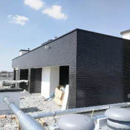 Budynek z elewacją z ciemnej, błyszczącej cegły na dachu, widoczne instalacje wentylacyjne i otoczenie placu budowy.