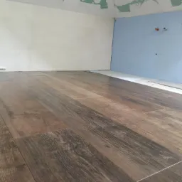 Ułożona podłoga z brązowych płytek imitujących drewno, widoczne ściany w trakcie wykończenia, częściowo zagruntowane, częściowo w kolorze jasnoniebieskim z wystającymi przewodami.