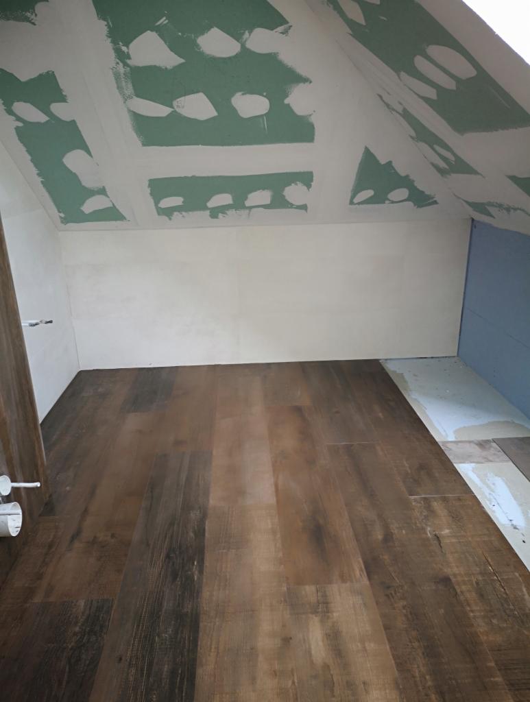 Podłoga z paneli imitujących drewno w nowym pomieszczeniu na poddaszu, ściany częściowo wykończone gładzią, sufit z widocznymi łączeniami płyt kartonowo-gipsowych przed szpachlowaniem.