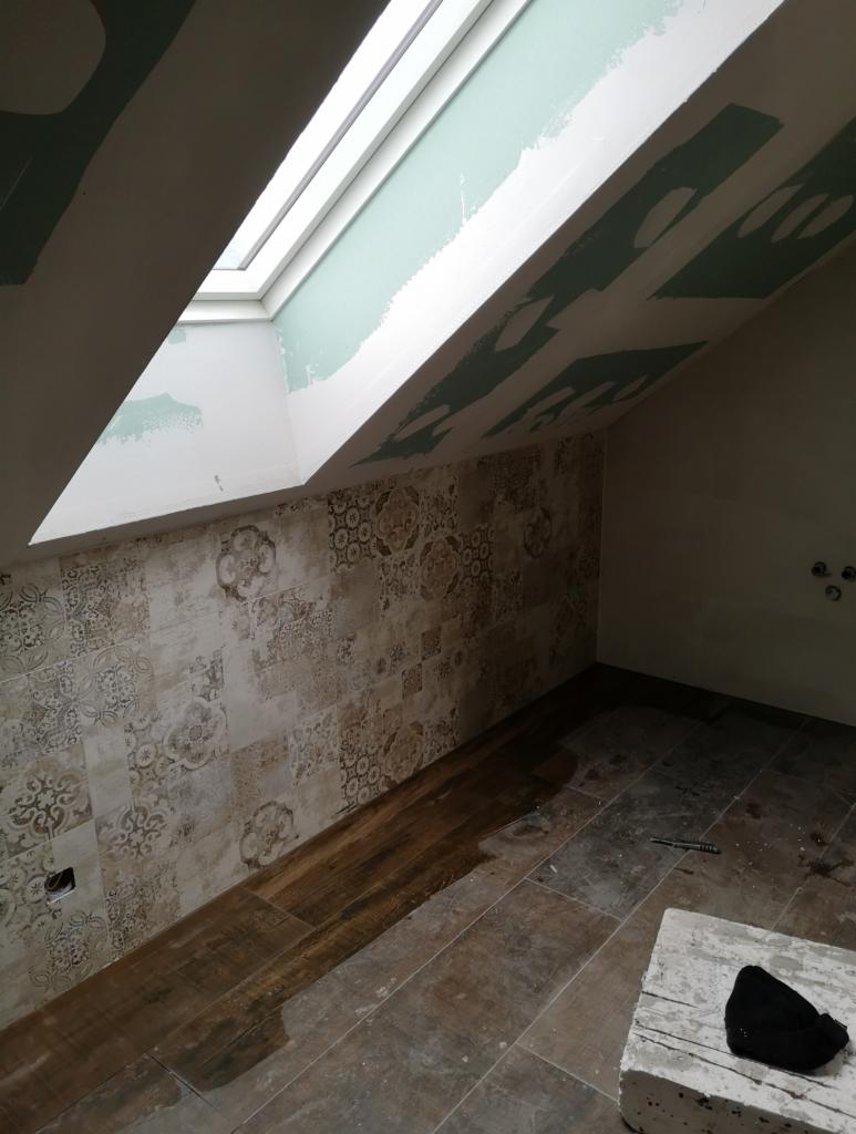 Ukończona łazienka na poddaszu z drewnopodobną podłogą z płytek i wzorzystą ścianą z płytek ceramicznych, widoczne okno dachowe i pozostałości po pracach wykończeniowych.