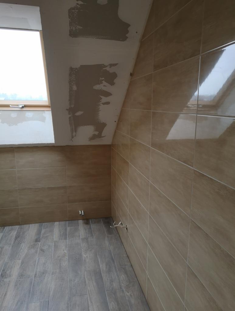 Wnętrze łazienki na poddaszu z beżowymi płytkami na ścianach i szarymi płytkami imitującymi drewno na podłodze, widoczne niedokończone prace wykończeniowe na suficie i rurki wystające ze ściany.