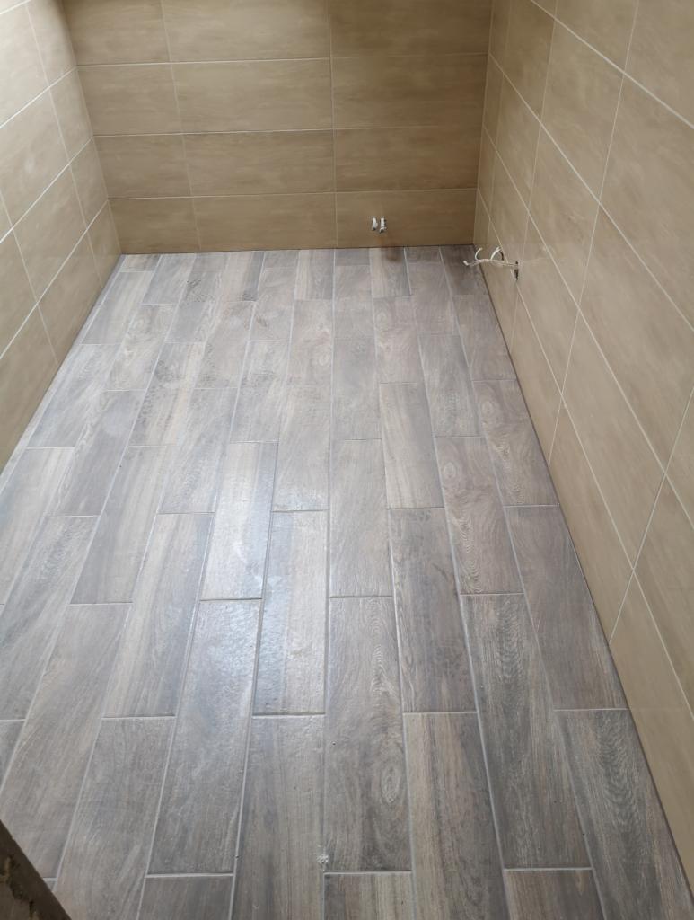 Wykończona łazienka z beżowymi płytkami ściennymi i podłogą z szarych płytek imitujących drewno, widoczne wystające kable instalacji.