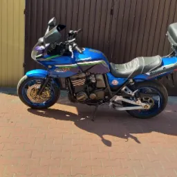 Niebieski motocykl Kawasaki ZRX 1200 z kufrem, zaparkowany na kostce brukowej, z brązowymi drzwiami garażowymi w tle i srebrnym autem osobowym.