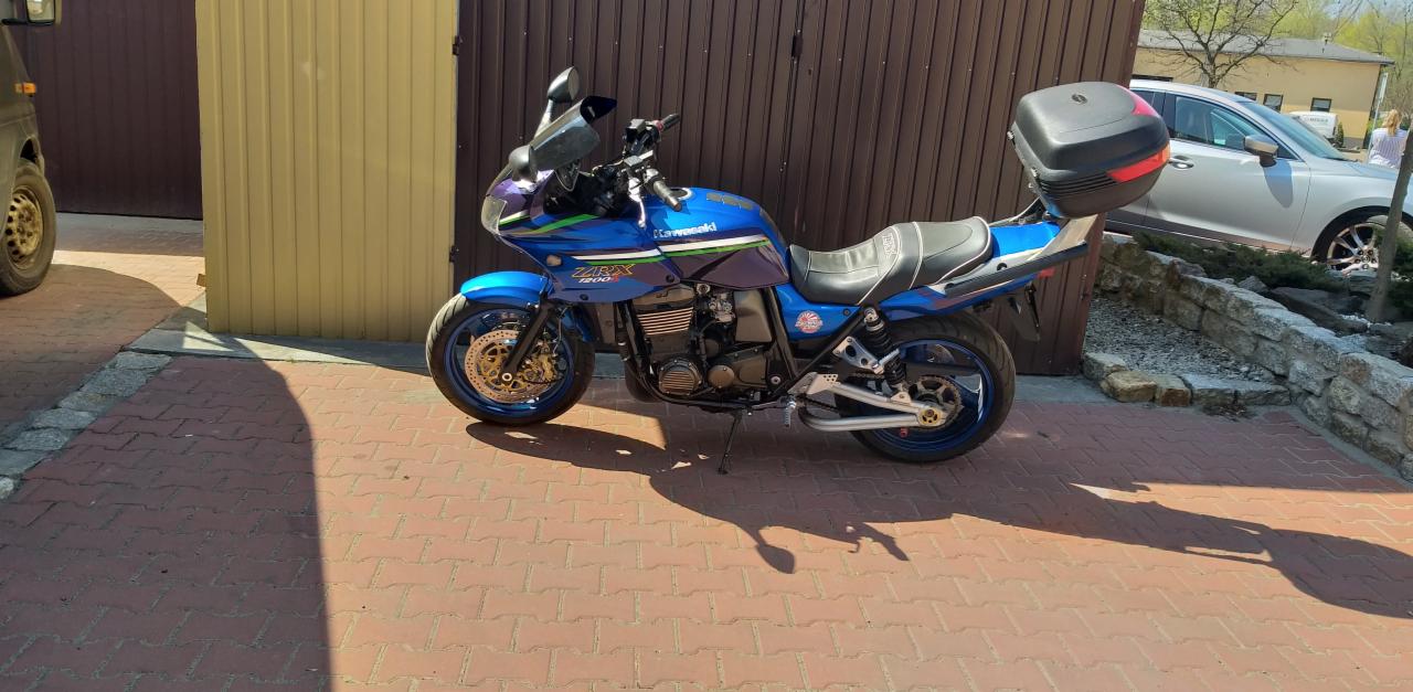 Niebieski motocykl Kawasaki ZRX 1200 z kufrem, zaparkowany na kostce brukowej, z brązowymi drzwiami garażowymi w tle i srebrnym autem osobowym.