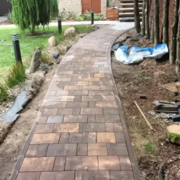 Kręta ścieżka z brązowej kostki brukowej prowadzi przez ogród. Widoczne drzewa, trawnik i elementy architektury domu w tle. Częściowo widoczne narzędzia.