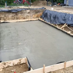 Zalany fundament pod budynek, zabezpieczony folią i szalunkiem z desek. Widoczny wykop i sprzęt budowlany w tle. Świeży beton.