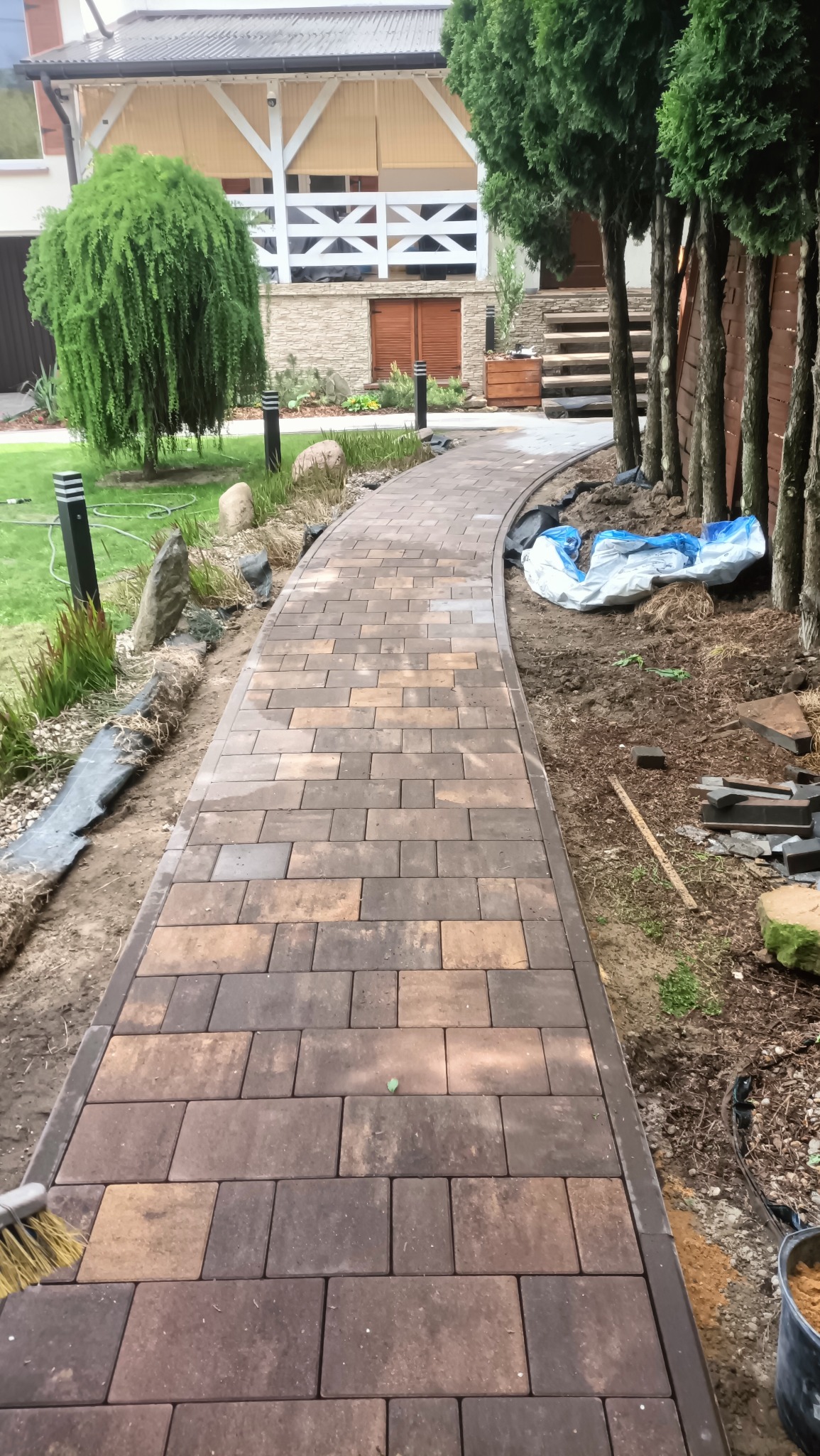 Kręta ścieżka z brązowej kostki brukowej prowadzi przez ogród. Widoczne drzewa, trawnik i elementy architektury domu w tle. Częściowo widoczne narzędzia.