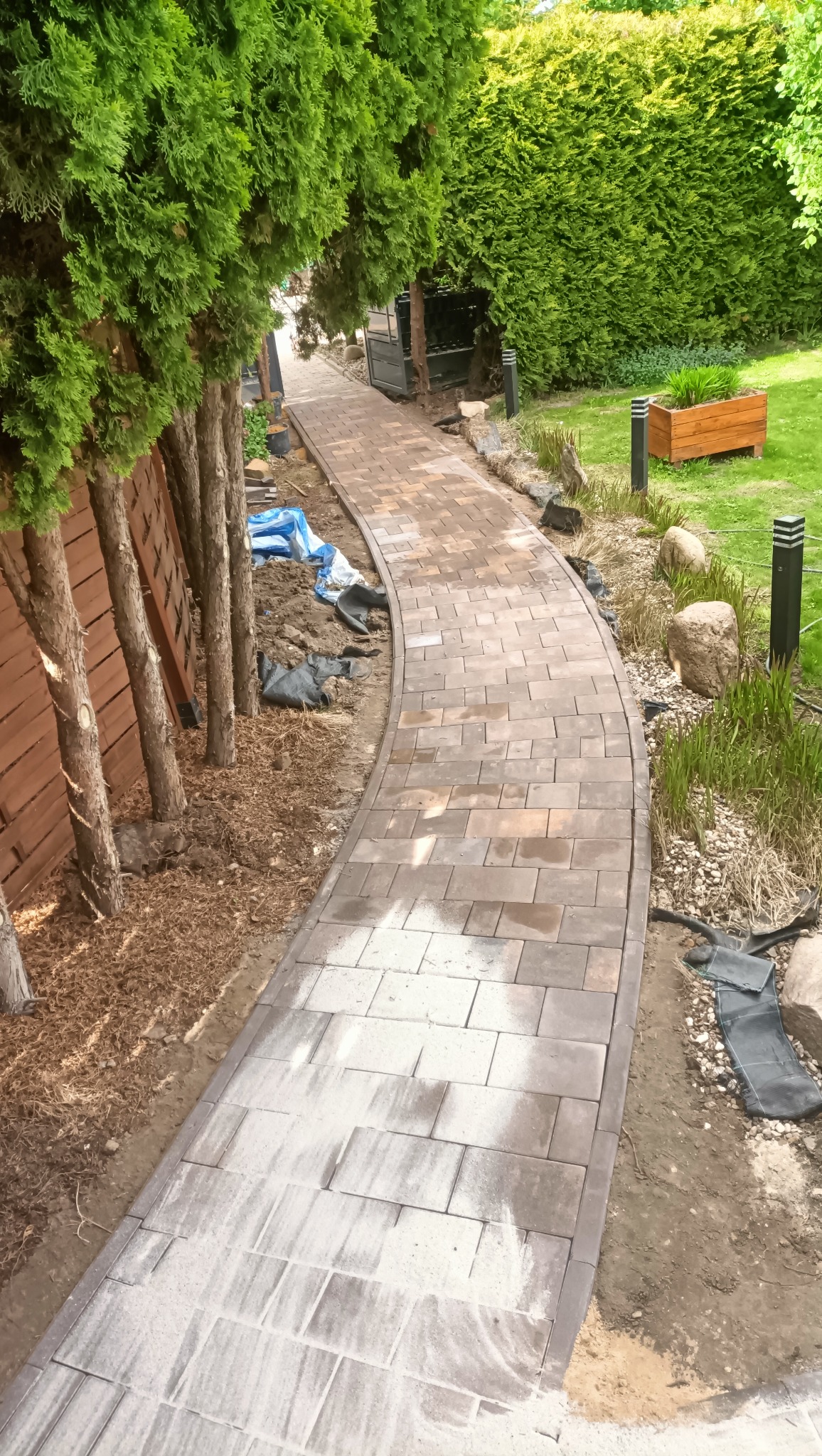 Kręta ścieżka z kostki brukowej w ogrodzie, widok z góry. Częściowo wykończona, z widocznymi materiałami budowlanymi i roślinnością w tle.