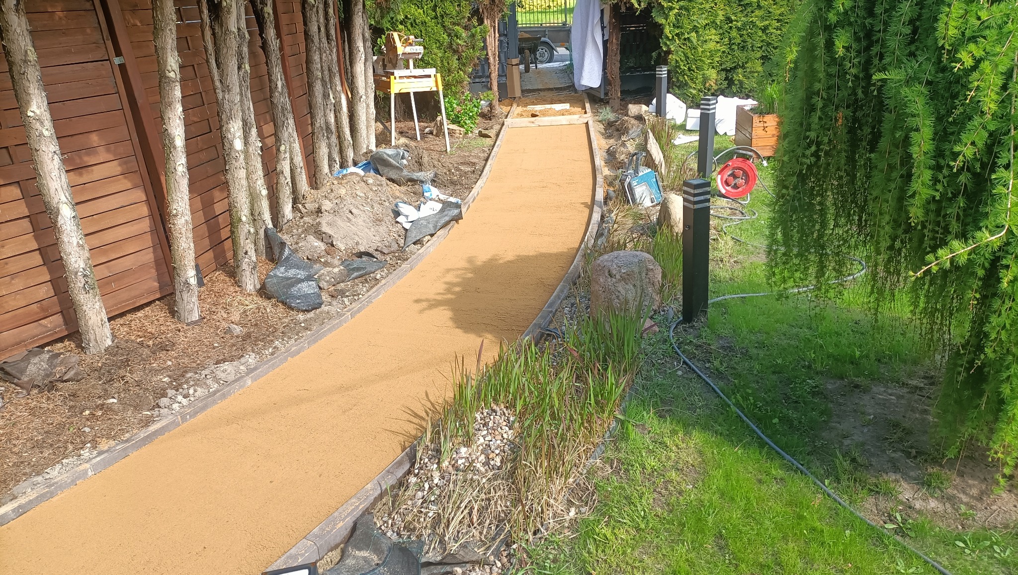 Ścieżka z piasku kwarcowego w ogrodzie, obrzeża z kostki, widoczne rośliny i drzewa. Etap prac brukarskich, przygotowanie podłoża, narzędzia w tle.