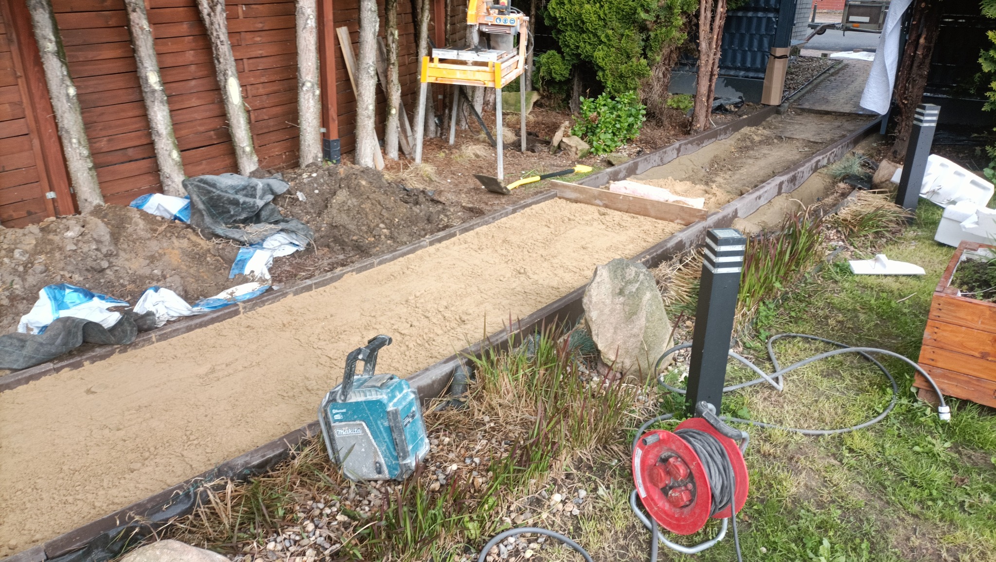 Przygotowanie podłoża pod ścieżkę w ogrodzie: piasek wyrównany w korycie z obrzeżami, widoczne narzędzia i materiały budowlane, lampy ogrodowe w tle.