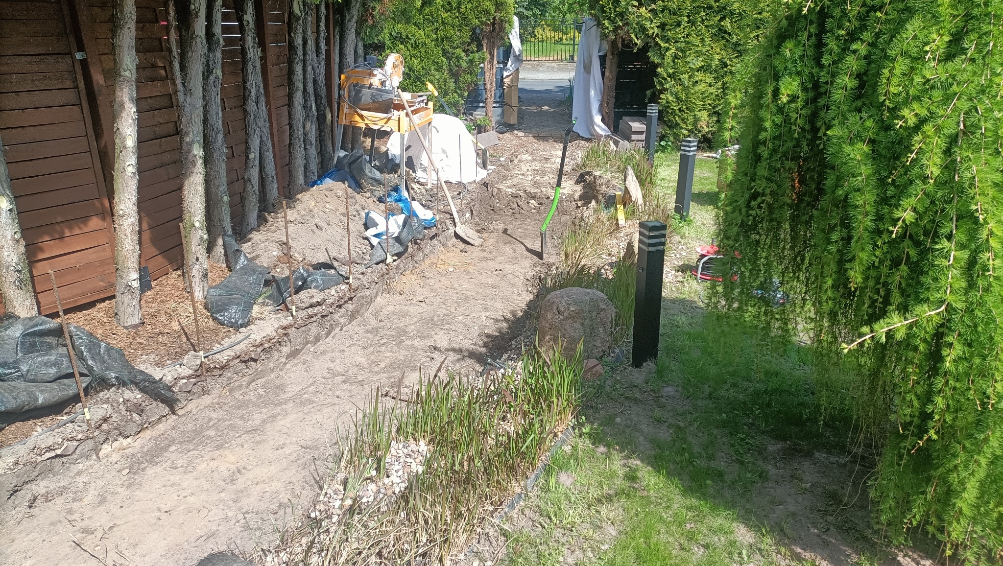 Przygotowanie terenu pod ścieżkę w ogrodzie: wykop, narzędzia, materiały budowlane. W tle widoczny mikser do betonu i elementy ogrodzenia. Słoneczny dzień.