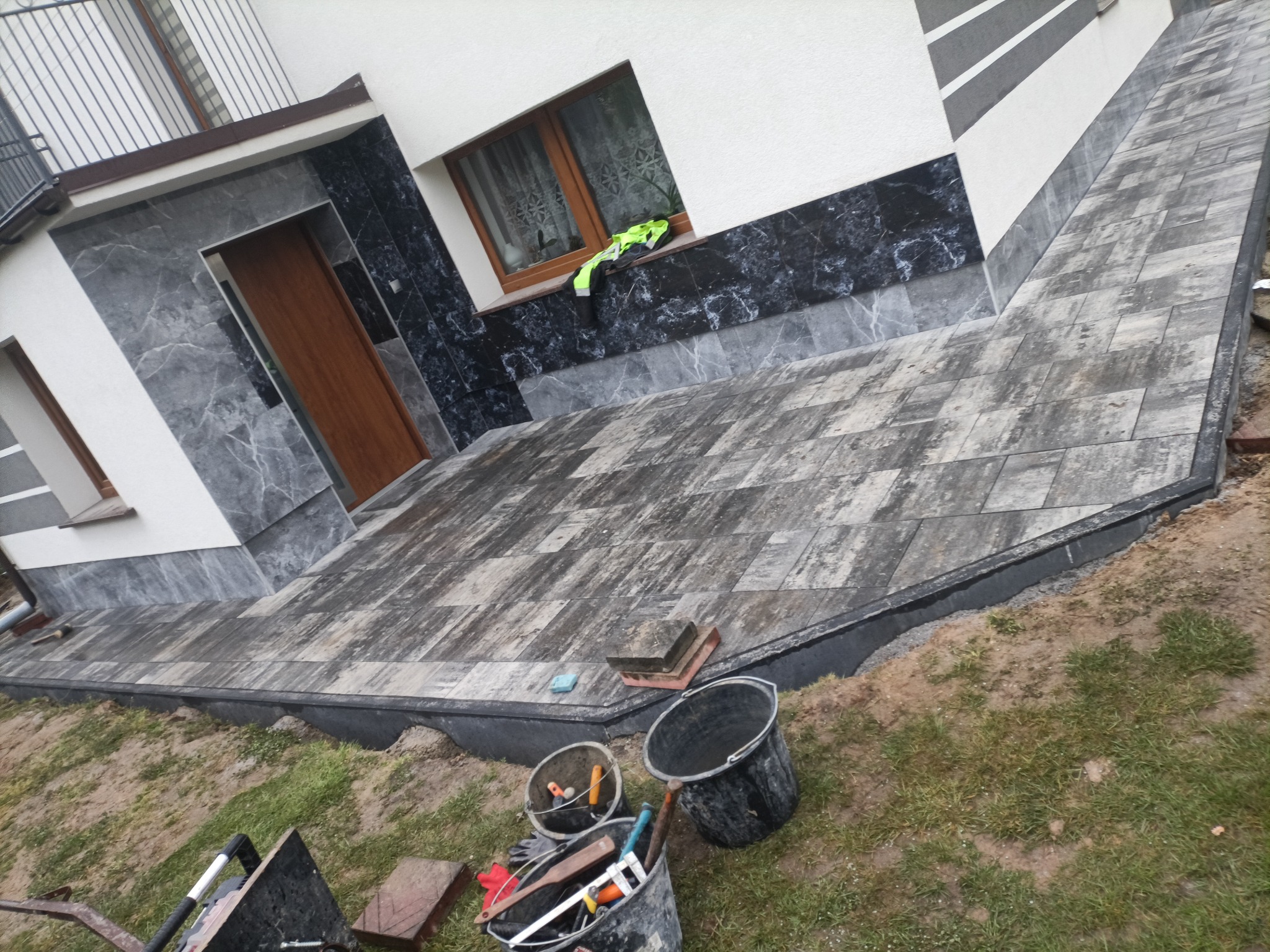 Ułożona kostka brukowa przed domem w Mokrsku. Widoczny fragment elewacji z oknem i narzędzia brukarskie. Kąt ujęcia z dołu, podkreślający wzór kostki.