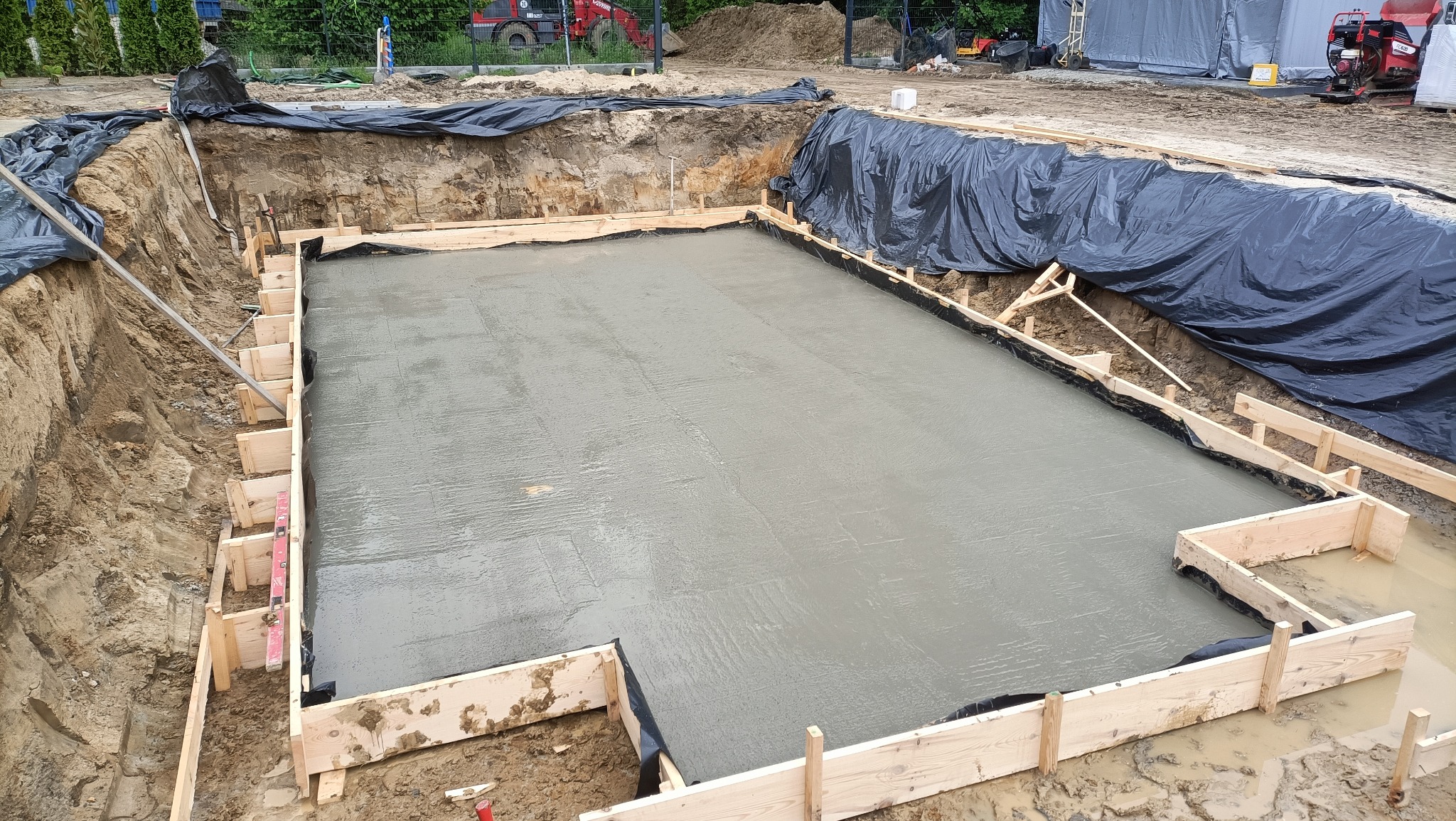 Zalany fundament pod budynek, zabezpieczony folią i szalunkiem z desek. Widoczny wykop i sprzęt budowlany w tle. Świeży beton.