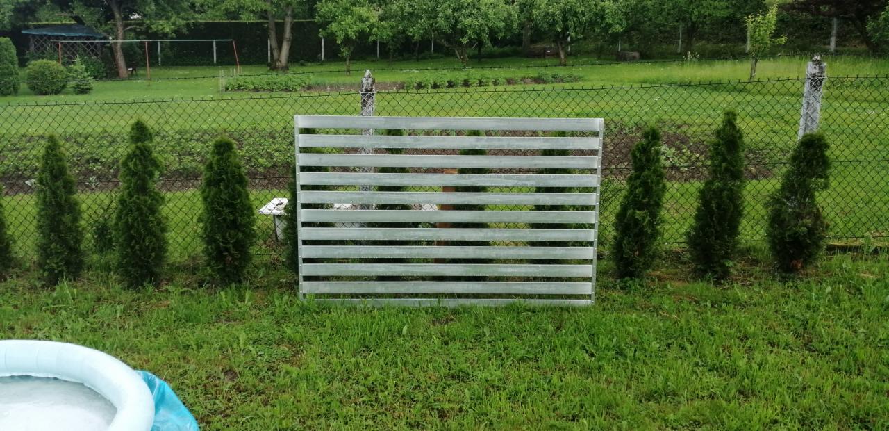 Panel ogrodzeniowy z poziomych, metalowych profili, ustawiony na trawie przed istniejącym ogrodzeniem z siatki, otoczony młodymi tujami; w tle drzewa i fragment basenu.